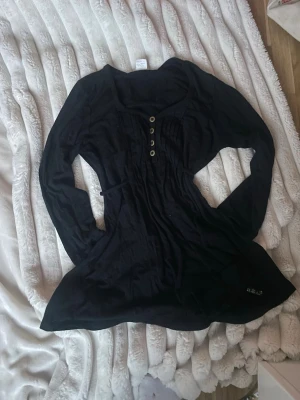 2000s topp - Svart långärmad blus med v-ringning och dekorativa knappar framtill. Modellen har en lös passform och lätt utställd nederdel. Perfekt för dig som gillar en enkel men ändå snygg stil. Det finns även band som man kan tajta den med som sitter på blusen,