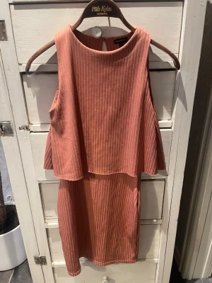 Rostfärgad ribbad kortklänning - Säljer en snygg rostfärgad kortklänning med ribbad struktur och ärmlös design. Klänningen har ett lager över bysten som ger en cool och modern look. Perfekt för dig som gillar stilrena detaljer och en avslappnad passform.