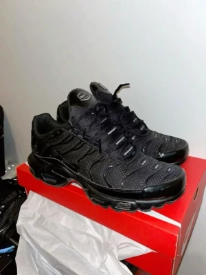 Nike Air Max Plus helsvarta sneakers - Nike Air Max Plus sneakers i helsvart med mesh- och syntetmaterial. Skorna har tydliga vågformade detaljer, synlig Air-dämpning i sulan och klassisk logga på plösen. Snygg och sportig siluett som passar till streetwear.