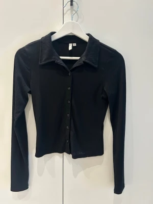 Svart ribbad topp med knappar - Stilren svart topp med ribbad struktur och långa ärmar. Tröjan har klassisk krage och knäppning framtill med svarta knappar. 