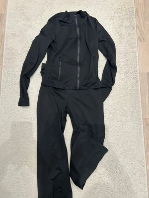 Svart träningsset med dragkedja - Säljer ett svart träningsset med långärmad jacka och byxor. Jackan har hel dragkedja framtill och två fickor med dragkedja. Setet är gjort i ett stretchigt och lätt material som passar perfekt för träning eller uppvärmning.