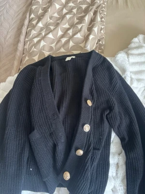 Svart stickad kofta från H&M - Snygg svart stickad kofta från H&M med grov ribbstickning och fyra dekorativa guldfärgade knappar framtill. Koftan har en djup V-ringning och långa ärmar, perfekt för lager på lager och en chill look.