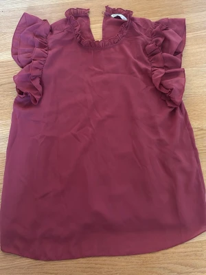 Vinröd blus med volang - Supersöt vinröd blus från ONLY med volangdetaljer vid ärmar och halsringning. Blusen är gjord i ett tunt, lätt tyg som känns luftigt och har korta ärmar med volanger för en trendig look. Perfekt att styla med jeans eller kjol.