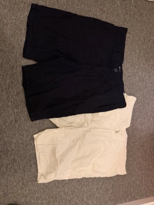 Vita och marinblå linneshorts herr - Två par shorts i klassisk modell, ett par vita och ett par marinblå. Båda är tillverkade i linne och har normal passform med knapp och dragkedja framtill. Perfekta för varma sommardagar och enkel att matcha med olika överdelar.