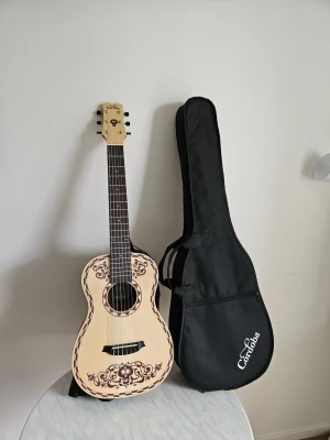 Cordoba Coco Mini akustisk gitarr - Säljer en Cordoba Coco Mini akustisk gitarr med ljus träfinish och dekorativa bruna mönster inspirerade av filmen Coco. Medföljer svart gigbag med Cordoba-logga. Perfekt för dig som gillar musik och snygg design.