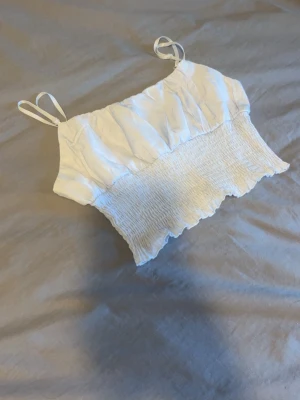 Vit croppad linnetopp med smock - Säljer en vit croppad linnetopp med smockad nederdel och tunna axelband. Toppen har en rynkad överdel och volangkant längst ner, perfekt för dig som gillar en söt och luftig stil. Materialet känns lätt och somrigt.