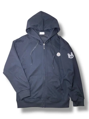 Svart Moncler hoodie med dragkedja - Svart hoodie från Moncler med dragkedja framtill, huva med snören och två fickor. Ikonisk Moncler-logga på bröstet och ett vitt broderat M-märke på ärmen. Tillverkad i mjukt bomullsmaterial som känns skönt mot huden. Perfekt för en chill och stilren look. INTE ÄKTA!
