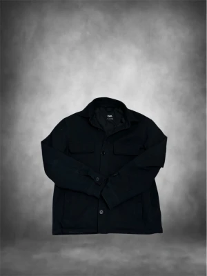 Svart overshirt från Zara - Stilren svart overshirt från Zara med klassisk krage, två bröstfickor med lock och knäppning framtill. Overshirten har raka ärmar och är tillverkad i ett mjukt tyg som ger en avslappnad look. Perfekt att bära över en t-shirt för en trendig lager-på-lager stil. Lite tjockare men en liten fläck som säkert går att få bort på bild två. Pris kan diskuteras 