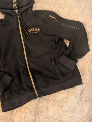 Svart Hugo Boss zip hoodie  - Svart hoodie från Hugo Boss med gulddetaljer och dragkedja med reflex framtill, trasig vid armarna och hål vid handleden annars inga problem, sällsynt