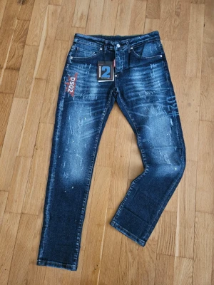 Mörkblå Dsquared2 jeans med slitningar - Snygga mörkblå jeans från Dsquared2 med coola slitningar och tvättade detaljer. Modellen har raka ben, klassisk femficksdesign och tydliga Dsquared2-loggor på både fram- och baksida. Perfekta för dig som gillar streetstyle och trendiga detaljer. Helt nya med prislapp kvar 