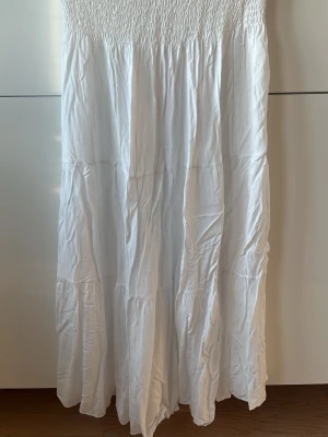 Vit långkjol i bomull - Luftig och flowy vit långkjol med smock i midjan och flera volanger. Kjolen är gjord i mjuk bomull och har en avslappnad passform som ger en skön och somrig vibe.