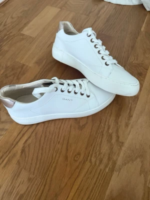 Vita sneakers från Gant i skinn - Stilrena vita sneakers från Gant i slätt skinn med vita snören och metallfärgade detaljer vid hälen. Klassisk låg modell med diskret logga på sidan och bekväm innersula. Perfekta för en clean och modern look.