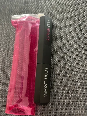 Huda Beauty Legit Lashes Mascara - Dubbeländad svart mascara från Huda Beauty med volym- och längd/lyft-borste. Kommer i en matt svart hylsa med vita och rosa detaljer samt en rosa genomskinlig förvaringspåse. Perfekt för att skapa dramatiska fransar.