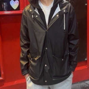 Svart regnjacka med huva - Snygg svart regnjacka. Jackan har tryckknappar framtill, två stora fickor och en rak passform. Perfekt för regniga dagar och enkel att matcha med olika outfits.