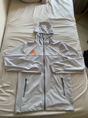 Ljusgrå vindjacka från The North Face - Ljusgrå vindjacka med huva från The North Face. Jackan har två dragkedjeförsedda fickor, elastiska muddar och en orange logotyp på bröstet. Materialet är lätt och passar perfekt för blåsiga dagar. Snygg och sportig design med diskreta detaljer.