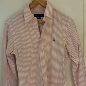 Rosa randig skjorta från Ralph Lauren - Säljer en klassisk rosa och vit randig skjorta från Ralph Lauren i slim fit-modell. Skjortan har långa ärmar, knappar framtill och den ikoniska broderade loggan på bröstet. Tillverkad i mjuk bomull och passar perfekt till jeans.