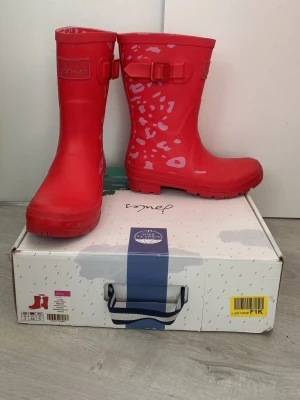 Röda gummistövlar från Joules - Snygga röda gummistövlar från Joules med ljusrosa leopardmönster och spänne på sidan. Tillverkade i slitstarkt gummi med grov sula för bra grepp. Perfekta för regniga dagar och festivaler.