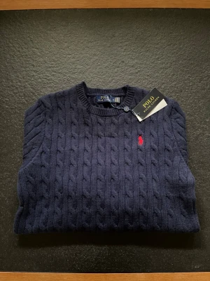 Sweater Polo Ralph Lauren Cable Knit 🧥 - Navy blue cable knit sweater by Polo Ralph Lauren with tags attached. Features the iconic red polo player logo.  ⭐️ Condition: 10/10 🎲 Size: M 🎨 Color: Navy Blue 📦 Fast and careful shipping