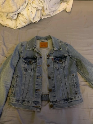 Ljusblå jeansjacka från Levi's - Klassisk ljusblå jeansjacka från Levi's med knappstängning framtill, två bröstfickor med lock och knappar samt två sidofickor. Jackan har en rak passform och typisk jeanskrage. Perfekt för en avslappnad och trendig look.
