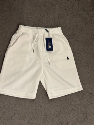 Ralph Lauren shorts - Säljer ett par vita shorts från Polo Ralph Lauren med elastisk midja och snörning. Klassisk logga broderad i svart på vänster ben. Sköna och stilrena shorts i mjukt material, perfekta för sommaren.