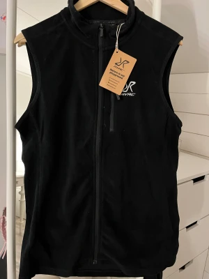 Svart fleeceväst från RVRC - Svart fleeceväst från RVRC med hög krage och hel dragkedja framtill. Västen har en stilren design med diskret logga på bröstet och är perfekt för lager-på-lager. Mjuk och skön fleece som håller värmen under kyliga dagar. Nu med prislapp, aldrig använd. 