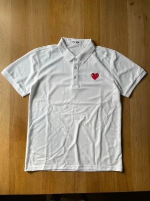 Vit pikétopp från Comme des Garçons Play - Vit pikétopp från Comme des Garçons Play med klassisk krage, tre knappar framtill och det ikoniska röda hjärte-emblemet broderat på bröstet. Tillverkad i mjuk bomull som känns skön mot huden. Perfekt för en clean och stilren look.