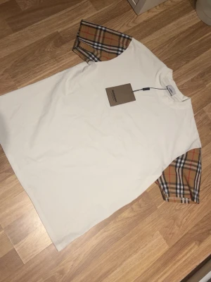 Vit Burberry t-shirt med rutiga ärmar - Säljer en clean vit t-shirt från Burberry med klassiska beige-rutiga korta ärmar. Oversized fit och rund halsringning. Perfekt för dig som vill ha en stilren men ändå unik look. Gjord i mjuk bomull för maximal komfort.helt ny aldrig använt 