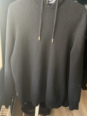 Svart kashmir/merinoull hoodie - En stilren svart hoodie med huva och dragsko. Hoodien har långa ärmar och ribbade muddar vid ärmslut och nederkant. Tillverkad i ett mjukt, stickat material som känns skönt mot huden. Perfekt för en avslappnad look.