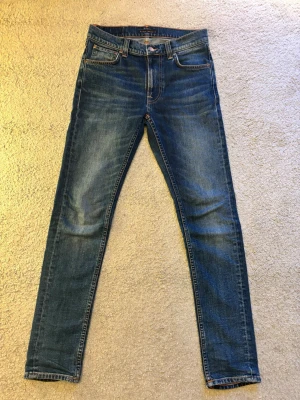 Blå slim fit jeans från Nudie (grim tim) - Snygga blå slim fit jeans från Nudie med klassisk femficksdesign och orangea sömmar. Jeansen har en något tvättad look och är tillverkade i denim med stretch för extra komfort. Perfekta för en avslappnad och trendig stil. Nypris är 1600 kr mitt pris 500 kr. Vid några frågor är det bara att höra av er !
