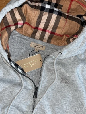 Grå Burberry zip hoodie - Snygg grå zip hoodie från Burberry med klassiskt rutigt foder i huvan och broderad logga på bröstet. Hoodien har dragsko i huvan, fickor framtill och ribbade muddar. Perfekt för dig som vill ha en stilren och exklusiv look.