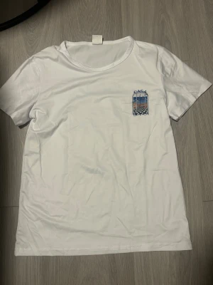 Vit t-shirt från Casablanca - Vit t-shirt från Casablanca med färgglatt tryck på ryggen och mindre tryck på bröstet. Motivet föreställer en utsikt genom ett fönster med texten 'Casa Blanca'. Klassisk passform och rund hals. Perfekt för en stilren och trendig look. Storlek M men passar mer som S