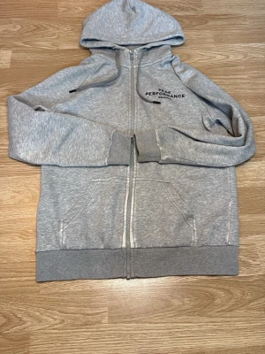 Grå Peak Performance hoodie med dragkedja - Ljusgrå hoodie från Peak Performance med dragkedja framtill, huva och två fickor. Broderad logga på bröstet. I storlek M.