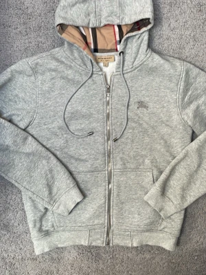 Grå zip hoodie från Burberry - Snygg grå hoodie från Burberry med dragkedja och klassiskt rutigt foder i huvan. Hoodien har dragsko, broderad logga på bröstet och är fodrad med mjukt material på insidan. Perfekt för dig som gillar exklusiv streetwear och vill ha något riktigt bekvämt.