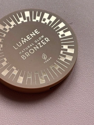 Lumene Natural Glow Bronzer - Lumene Natural Glow Bronzer i en rund, brun plastförpackning med silvriga detaljer. Pudret har en ljusbrun, naturlig nyans och en pressad mönstrad yta. Vegansk formula som ger en fräsch solkysst look.
