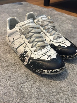 Maison Margiela svartvit sneakers - Unika sneakers från Maison Margiela i vitt läder med svart färgstänk-design. Klassisk låg modell med vita snören och coola detaljer som sticker ut. Perfekta för dig som vill ha något annorlunda och edgy i garderoben.