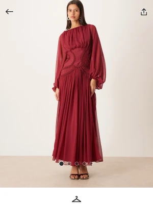 Vinröd långklänning med volangärm - Stilren vinröd långklänning med markerad midja och transparenta, långärmade volangärmar. Klänningen har ett svepande fall och fina detaljer vid midjan som ger en elegant siluett. Perfekt för dig som vill sticka ut.