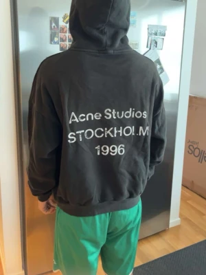 Svart hoodie Acne Studios Stockholm - Svart hoodie från Acne Studios med stor vit text på ryggen där det står 'Acne Studios STOCKHOLM 1996'. Klassisk passform med huva och ribbade muddar. Perfekt för dig som gillar streetwear och vill ha något från ett ikoniskt svenskt märke.