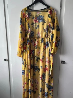Gul blommig långklänning med knyt - Säljer en långklänning i härlig gul färg med blommigt mönster och knyt i midjan. Klänningen har långa ärmar och ett flowigt fall som ger en avslappnad och snygg look. Perfekt för dig som vill sticka ut med färg och mönster!