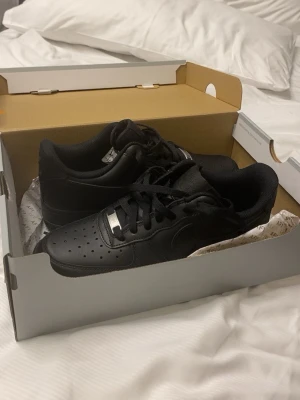 Svarta Nike Air Force 1 sneakers - Snygga nya helsvarta Nike Air Force 1 sneakers med klassisk låg siluett. Skorna har ovandel i slätt läder, perforerad tå för ventilation och svart yttersula. Ikonisk Nike-logga på sidan och plös. Perfekta för en stilren streetlook.