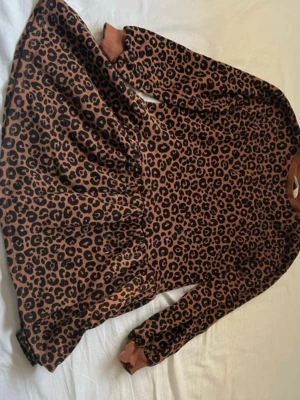 Leopardmönstrad klänning H&M barn - Leopardmönstrad kortklänning från H&M i bruna och svarta toner. Klänningen har lång ärm, ribbade muddar och är gjord i mjuk bomull. Perfekt för dig som gillar djurmönster och vill ha en bekväm men snygg look.