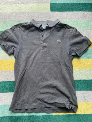 Grå pikétröja från Lacoste Sport - Snygg grå pikétröja från Lacoste Sport med klassisk krage och tre knappar framtill. Ikoniska gröna krokodilloggan broderad på bröstet. Perfekt för en sportig och stilren look. Tillverkad i mjukt bomullsmaterial.