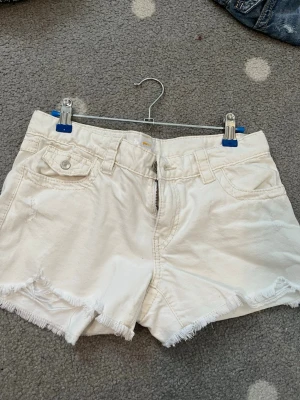 Vita fransiga jeansshorts - Säljer ett par vita jeansshorts med fransig kant och klassisk femficksdesign. Shortsen har dragkedja och knapp framtill samt bälteshällor. Perfekta för varma dagar och ger en avslappnad vibe.