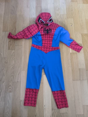 Spiderman dräkt med mask - Cool Spiderman-dräkt i blått och rött med svarta spindelnätsdetaljer och stor spindel på bröstet. Dräkten har långa ärmar och ben samt medföljande mask med vita ögon. Perfekt för cosplay eller maskerad!