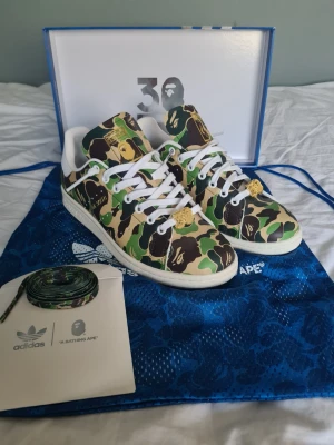 Adidas x BAPE Stan Smith camo sneakers - Unika sneakers från Adidas och BAPE i Stan Smith-modell med klassiskt grönt, brunt och beige camo-mönster. Vita detaljer, guldfärgade loggor på hälen och vita snören. Tillverkade i läder med ikoniska BAPE och Adidas-detaljer. Kommer med extra camo-skosnören och originalpåse.