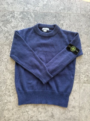 Blå stickad tröja från Stone Island - Mysig blå stickad tröja från Stone Island med klassisk rund halsringning och ribbade muddar. Tröjan har den ikoniska Stone Island-loggan på vänster ärm. Perfekt för lager-på-lager och chill stil.