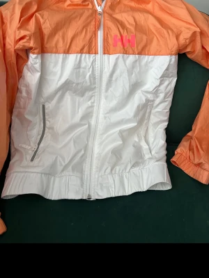Helly Hansen orangevit vindjacka - Snygg vindjacka från Helly Hansen i orange och vitt med dragkedja framtill och två fickor med dragkedja. Jackan har meshfoder för extra komfort och HH-logga i rosa på bröstet. Perfekt för blåsiga dagar och sportiga outfits.