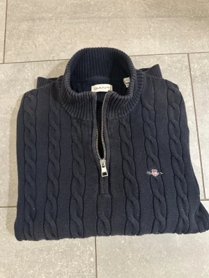 Mörkblå kabelstickad GANT half zip - Mörkblå kabelstickad tröja från GANT med halv dragkedja och ribbad hög krage. Klassisk broderad GANT-logga på bröstet. Perfekt för dig som gillar stilrena och tidlösa plagg. Passar XS-möjligen S
