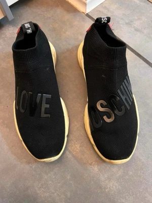 Svarta Moschino sneakers med text - Snygga svarta Moschini sneakers. Kolla bilder för skavanker, ena sulan är sönder under 