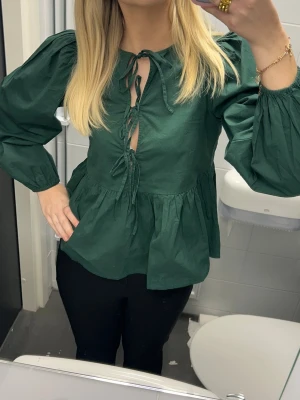 Grön blus med knytning framtill - Snygg mörkgrön blus med volang nertill och puffiga långärmar. Blusen har knytning framtill och en lätt v-ringning🩷