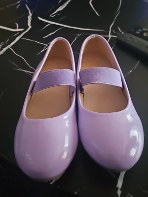 Ljuslila ballerinaskor med resårband - Snygga ballerinaskor i glansigt ljuslila material med matchande resårband över foten. Skorna har rund tå och låg sula, perfekt för en trendig och bekväm look. Innersulan är beige och materialet ser ut att vara syntetiskt.